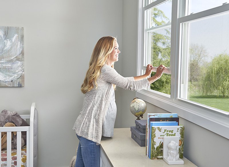 A smiling pregnant blonde woman in a nursery, reaching over a dresser to lower a window.
