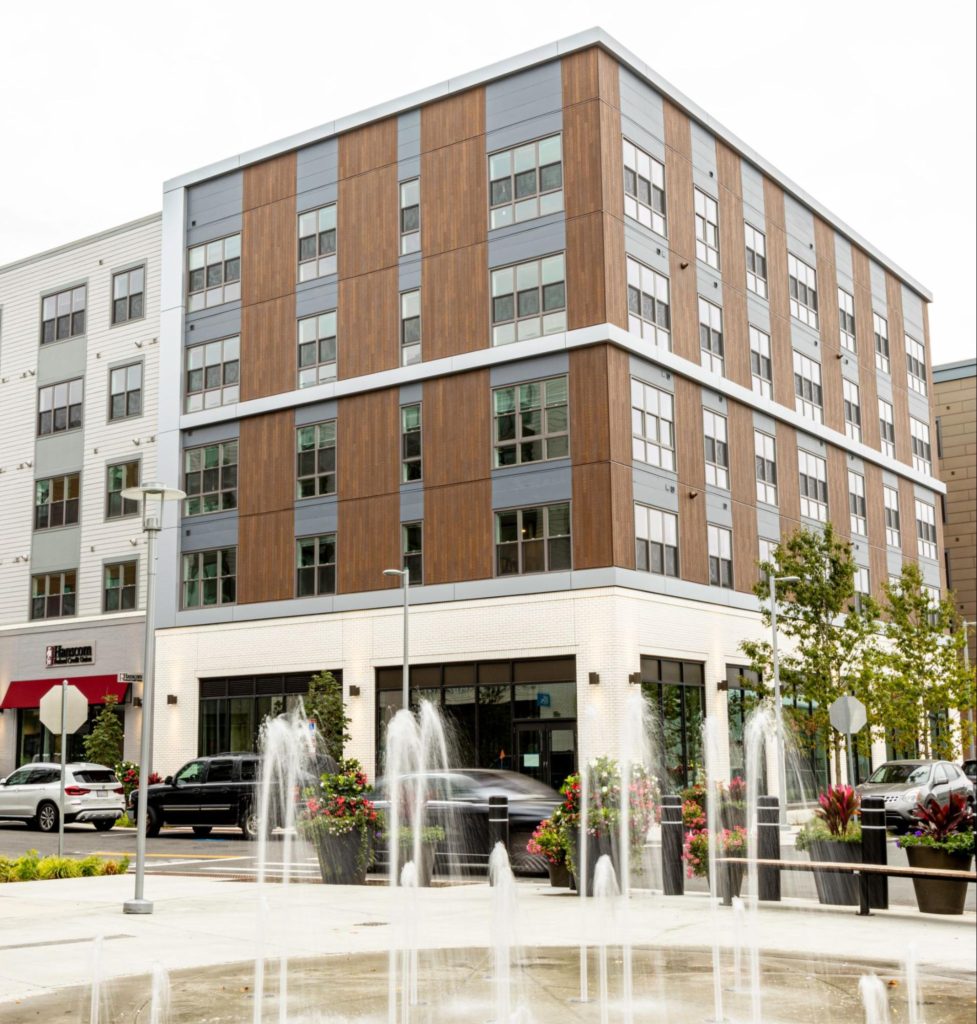 A view from behind a splash pad of the corner of a five-story commercial building with white brick on the bottom floor and vertical wood cladding on the top.