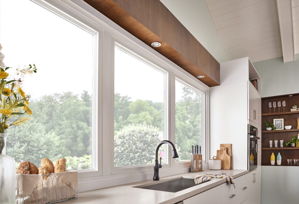 A clean white kitchen with three large windows behind a single-basin kitchen sink.