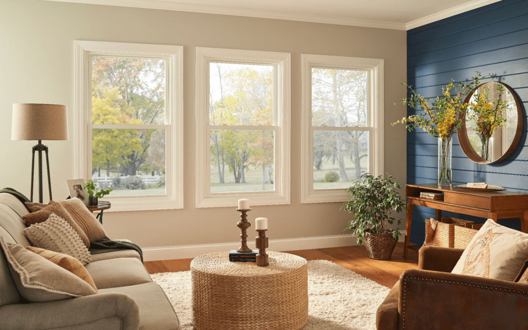 An interior view of a living room with three vinyl windows showing the fall trees outside, and a navy blue shiplap adjoining wall. There is a beige couch and armchair, and a brown desk