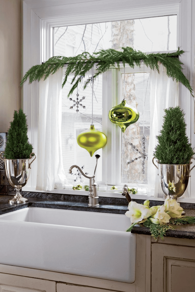 This kitchen design has a white farmhouse sink and a dark marble countertop. There are big ornaments hanging in front of the kitchen window, two small trees in silver vases on the sides of the sink and some garland draped across the window.
